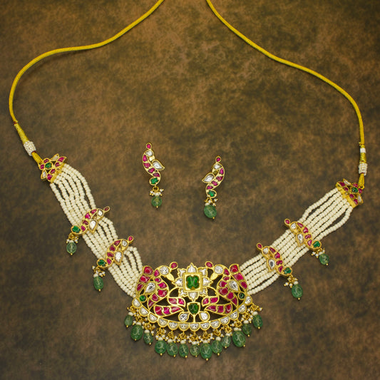 Jadau Kundan necklace set with intricate red and green gemstone detailing, multiple strands of white pearls, and matching earrings featuring emerald bead drops, set in pure 22k gold plating.