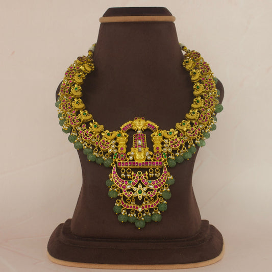 This is a short necklace with lord Balaji on it. The stones are jadau Kundan work and it’s 22k gold plated. At the bottom of the necklace there are Russian emerald beads