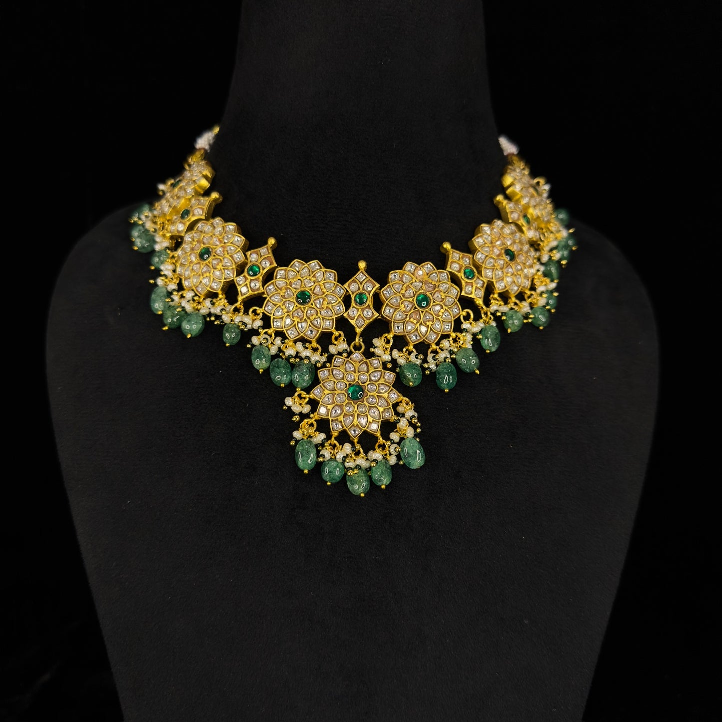 Here is A short Jadau Kundan bottumala necklace with floral design. This necklace has white Kundan stones and green stones in the middle of flowers. This piece is covered with 22k gold plating.