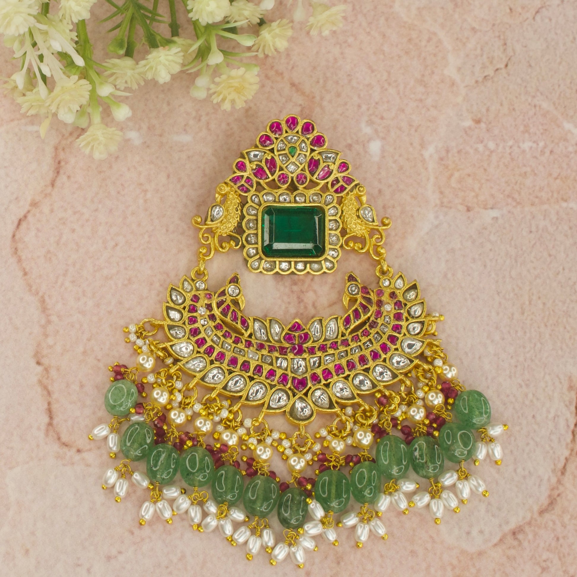 This is a jadau Kundan stone pendant with an emerald in middle. The pendant is large sized, it is covered with 22k Gold plating and at the bottom of the pendant we have strawberry beads and ricepearls
