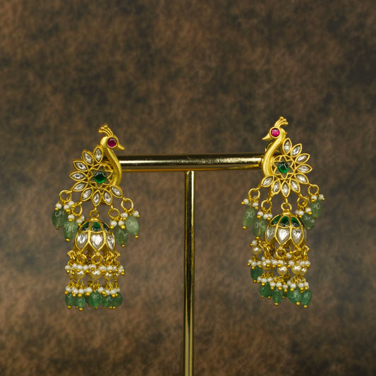 Jadau Kundan jhumkas with peacock motif, vibrant red and green gemstones, white Kundan stones, pearl drops, and emerald beads, set in pure gold plating.